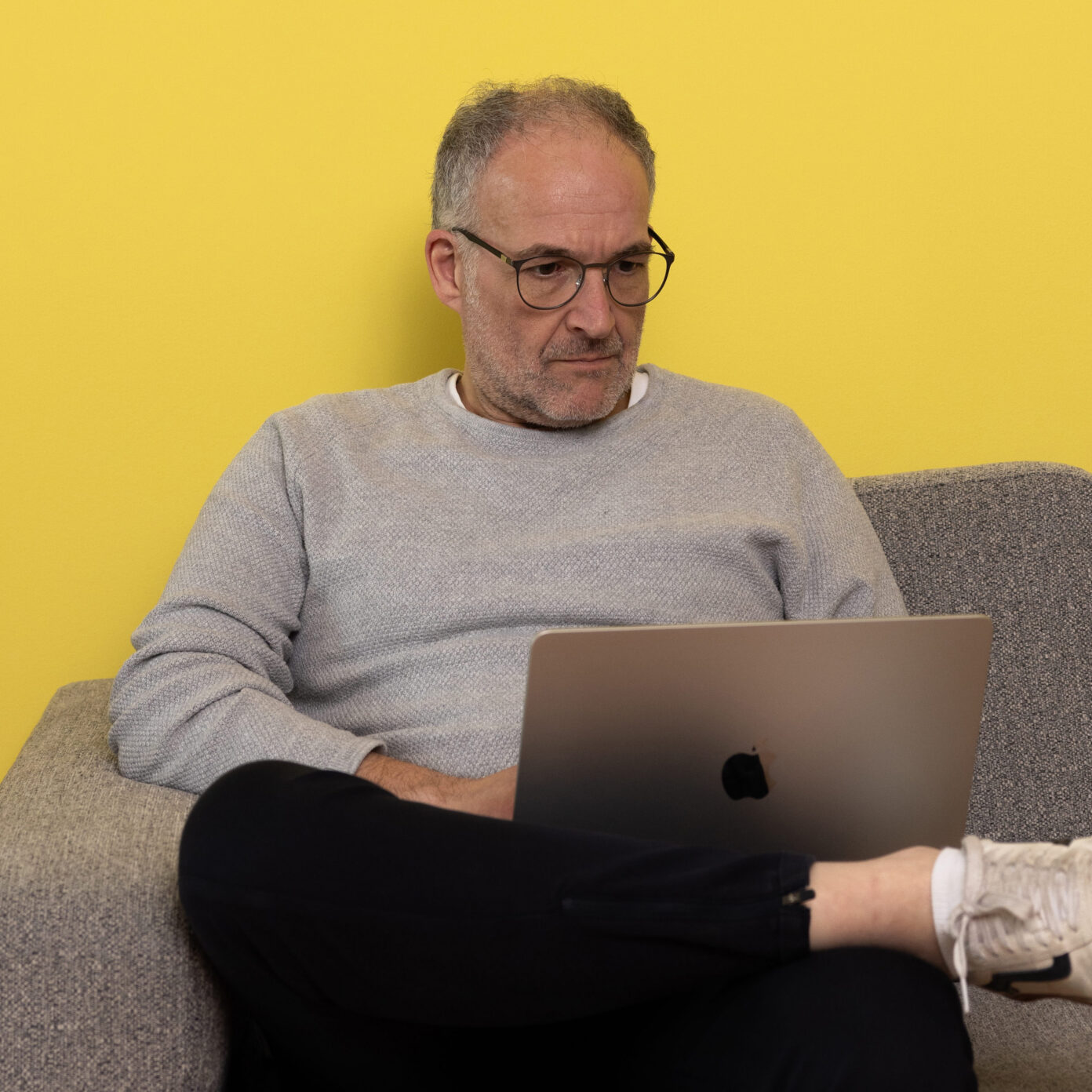 Foto von Mirko, Projektmanager bei der Digitalagentur seitenweise in Bielefeld. Mirko sitz auf einem grauen Sofa vor einer gelben Wand, hat einen Laptop auf dem Schoß und arbeitet konzentriert.